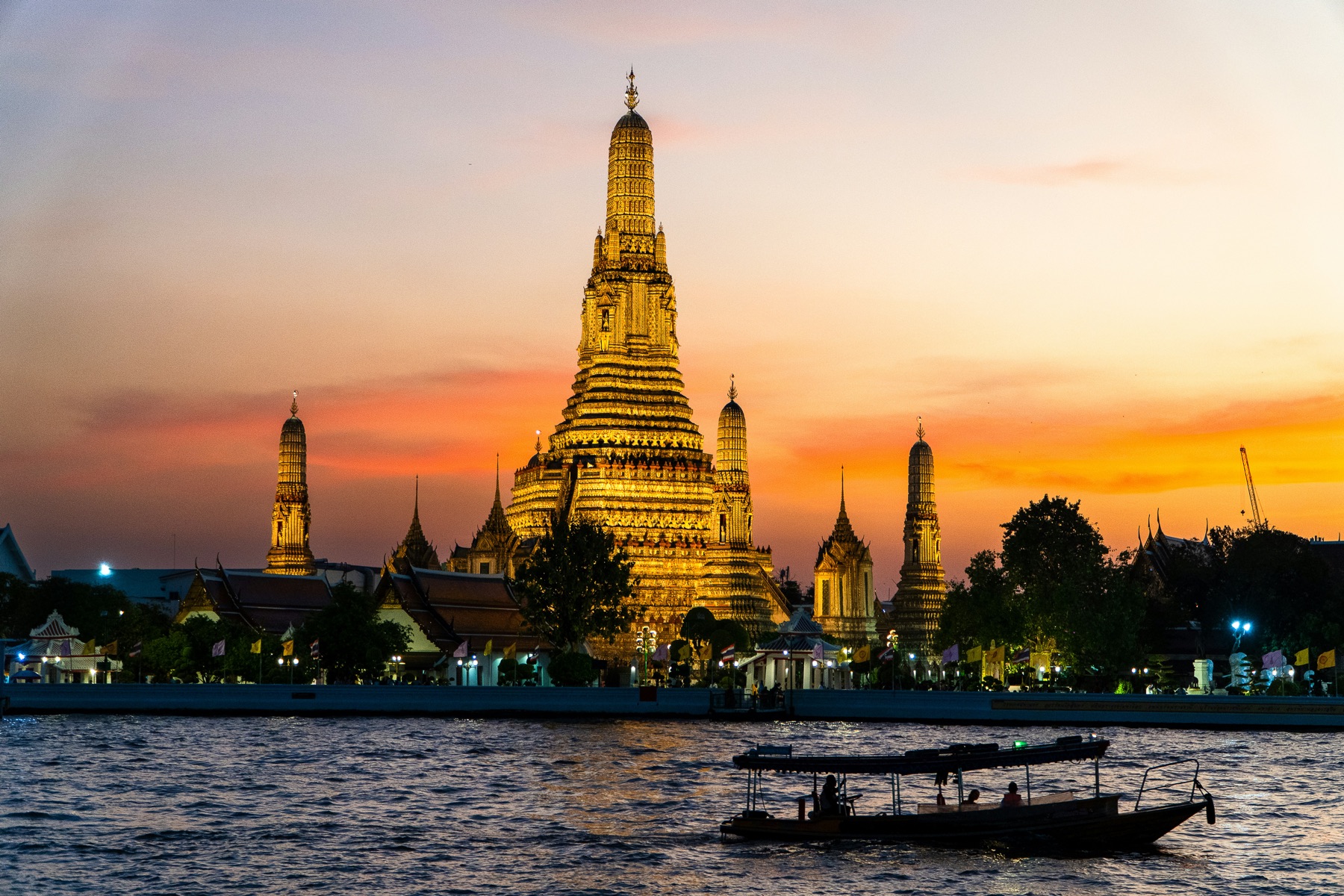 Osvetlený chrám Wat Arun pri západe slnka nad riekou Chao Phraya v Bangkoku, v popredí loď